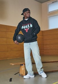 Felpa nera con il logo dei "Chicago Bulls", jeans azzurri chiari e sneakers bianche, mentre tiene in mano un pallone da basket nero, in piedi all'interno.
