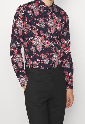 Mann trägt langärmliges schwarzes Hemd mit rotem und weißem Blumenmuster, in hoch taillierte schwarze Hose gesteckt, Hände teilweise in den Taschen.