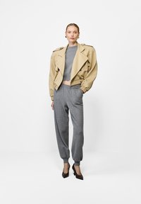 Naistele, kes kannab beežikat lühikest jakki halli sviidri ja sobivate hallide jogger-pükste kohal, jalas pruunid kontsakingad, seismas valge tausta ees.