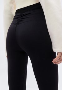 Μαύρο κολάν με ψηλή μέση και ρυτιδιασμένη λεπτομέρεια, κατασκευασμένο από ένα λείο, ελαστικό ύφασμα. Η εμφάνιση ολοκληρώνεται με ένα εκρού μακρυμάνικο τοπ.