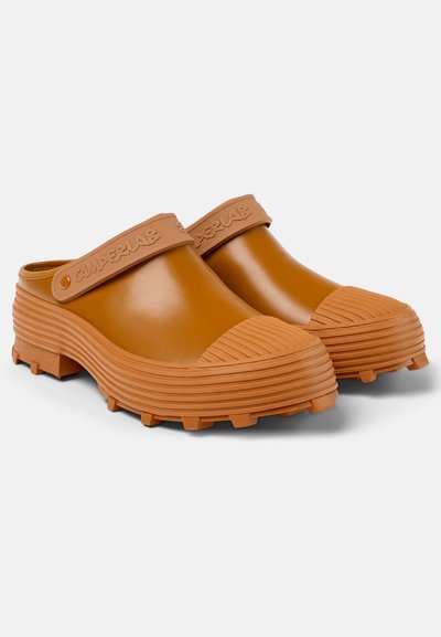 Rubber clogs in a warm brown color, featuring a ridged toe cap, textured strap with logo, and a chunky sole with traction studs.
