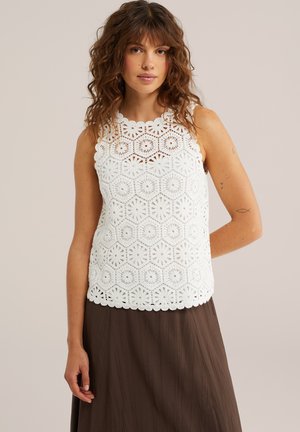 Žena s vlnitými hnedými vlasmi má na sebe biely háčkovaný top bez rukávov a hnedú plisovanú sukňu, stojaca pred jednoduchým béžovým pozadím.