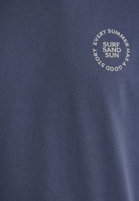 Navy blue cotton t-shirt with a circular white graphic text reading "SURF SAND SUN EVERY SUMMER HAS A GOOD STORY."