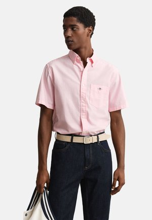 Mężczyzna ubrany w jasnoróżową, krótkim rękawem koszulę zapinaną na guziki, ciemne dżinsy, beżowy pasek, trzyma biało-granatową marynarkę w paski.