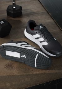 Chaussures de course Adidas noir et blanc placées sur un sol en bois avec deux haltères noires à proximité.