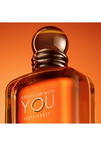 Bottiglia di profumo in vetro color ambra con tappo tondo in ambra scura, etichettata "Stronger With You Intensely" su sfondo arancione.