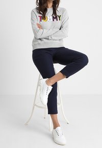 Sweatshirt gris avec "TOMMY" en lettres multicolores, associé à un pantalon cropped bleu marine foncé et des baskets blanches. Look élégant et décontracté.