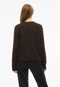 Brązowy sweter o teksturze z dekoltem w serek, z długimi rękawami i luźnym krojem, w połączeniu z czarnymi spodniami, pokazany od tyłu.