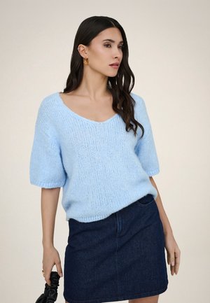 Mujer con cabello largo y oscuro que lleva pendientes dorados, un suéter de punto de manga corta azul claro, falda vaquera oscura y sostiene una bolsa scrunchie negra.
