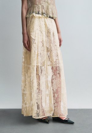 Parte inferior del cuerpo vistiendo una falda larga beige translúcida de encaje con patrón floral sobre pantalones cortos, combinada con zapatos planos negros puntiagudos.