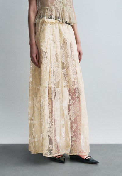 Bas du corps portant une longue jupe beige en dentelle transparente avec un motif floral sur un short, assortie à des ballerines noires pointues.