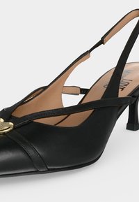 Μαύρο δερμάτινο γοβάκι slingback με χαμηλό τακούνι kitten, σταυρωτά λουράκια μπροστά και χρυσό στρογγυλό έμβλημα στο δάκτυλο.