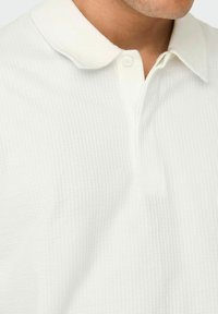 Hvid polo-shirt med tekstureret stof, blød krave, tre-knap front og et subtilt præget mønster.