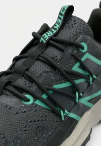 Chaussure de sport noire avec une tige texturée ornée d'accents mouchetés, des détails verts et des lacets noirs. La semelle est d'une couleur claire contrastante.