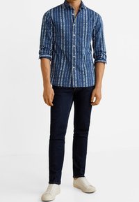 Camicia button-up blu a motivi con strisce verticali bianche, abbinata a jeans scuri e sneakers chiare. La camicia ha un colletto e maniche arrotolate.