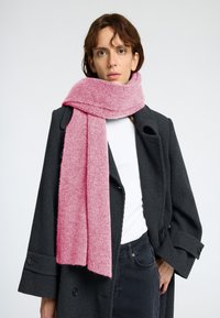 Mjuk, ribbad rosa halsduk hängande runt halsen, som kompletterar en mörk houndstooth-kappa. Har en lång, rektangulär form och varm textur.