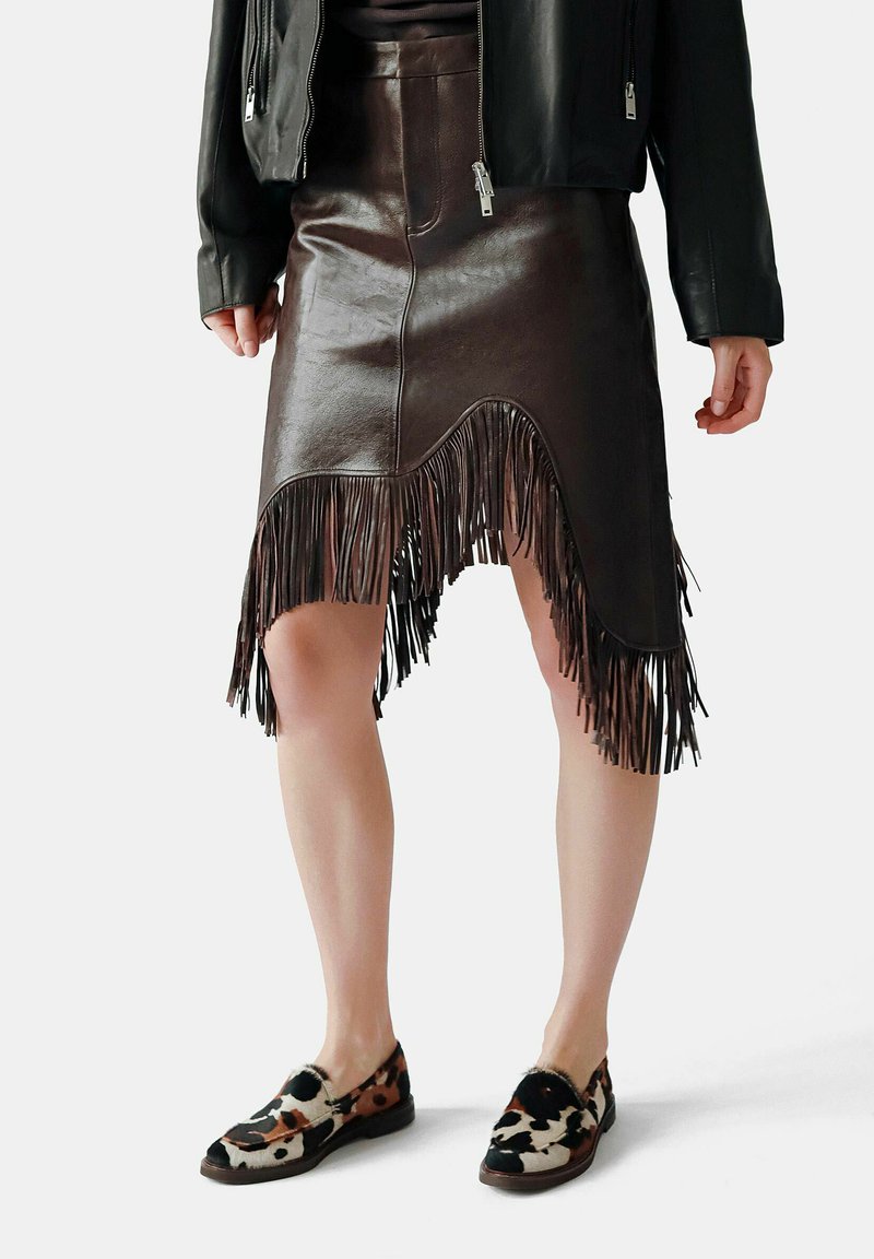 Jupe en cuir marron avec des détails en franges à l'ourlet, présentant une coupe ajustée et des zips. Portée avec des chaussures à enfiler à motifs.