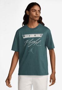 Teal bombaška majica s kratkimi rokavi, ki ima bel grafični napis "NIKE Flight" in sproščen kroj. Gladka tekstura brez vidnih vzorcev.