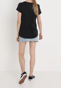 Frau mit langen Haaren, die ein schwarzes T-Shirt, einen hellblauen ausgefransten Denim-Minirock und schwarze Vans-Sneaker trägt, geht vor einem weißen Hintergrund.