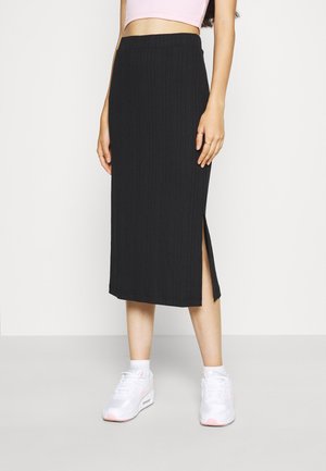 Jupe midi noire à côtes avec une fente sur le côté, présentant une coupe ajustée et une taille élastique. Associée à des baskets blanches.