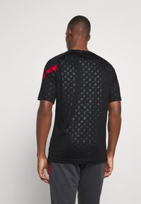 Černé sportovní tričko s krátkým rukávem s vyraženým vzorem, červenými akcenty na ramenou, lehká látka a rovný lem. Zobrazen pohled zezadu.