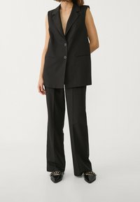 Musta räätälöity liivi, jossa syvä V-pääntie, isot kaulukset ja kaksi nappia. Yhdistetty mustiin, leveälahkeisiin housuihin ja teräväkärkisiin kenkiin.