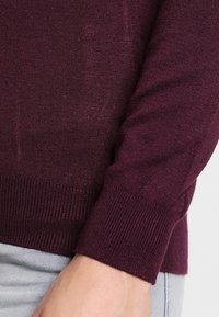 Bordowy sweter z dzianiny z prążkowanymi mankietami, gładką teksturą i dopasowanym krojem. Widoczna ręka trzymająca rąbek, w połączeniu z jasnoszarymi spodniami.