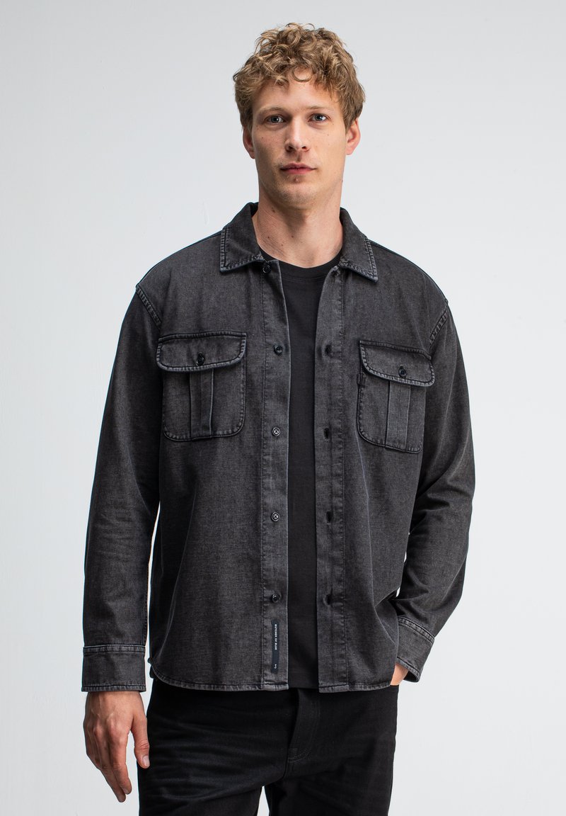 Grijze, lange mouwen shirt met button-down kraag, twee borstzakken en zwarte knopen. Gedragen over een zwart t-shirt, gemaakt van een denim-achtige stof.