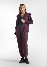 Giacca zip-up bordeaux con cappuccio, abbinata a pantaloni tapered coordinati. Il top presenta un colletto rotondo e una texture liscia. Le sneaker nere completano il look.