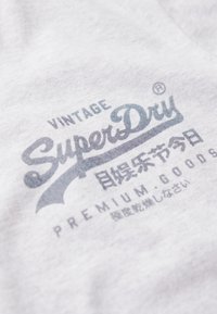 Grå bomulds t-shirt med et slidt blåt grafik. Teksten inkluderer "VINTAGE SuperDry" og "PREMIUM GOODS" i forskellige skrifttyper.
