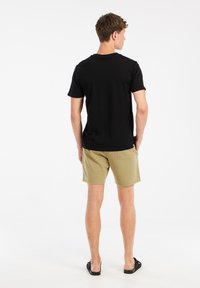 Zwarte T-shirt met korte mouwen, van katoen; gecombineerd met lichte khaki shorts en zwarte sandalen. Het zicht is van achteren, waarbij de outfit wordt benadrukt.