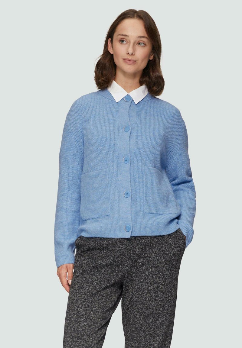 Hellblau gestrickte Strickjacke mit Knopfverschluss, zwei vorderen Taschen und einer entspannten Passform, kombiniert mit grauen gemusterten Hosen und einem weißen Kragenhemd.