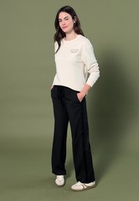 Woman standing with hands in pockets wearing white sweatshirt, black pants, and white sneakers against plain olive background.