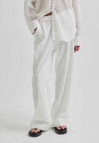Bílý outfit složený z volné košile s dlouhými rukávy a širokými kalhotami s mřížkovým vzorem. Oba kusy jsou vyrobeny z lehké, strukturované tkaniny.