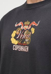 Zwart shirt met ronde hals en een kleurrijke afbeelding van een kroon, een rood lint, gouden munten en de tekst "Velvet Store Seal of Approval COPENHAGEN."