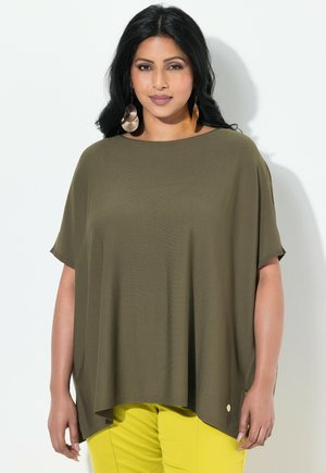Olivengrünes oversized Oberteil mit kurzen Ärmeln, Rundhalsausschnitt und seitlichem Schlitzdetail. Kombiniert mit hellgelben Hosen.