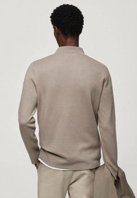 Hombre lleva una camisa de manga larga beige y pantalones a juego, sosteniendo una chaqueta beige, de pie con la espalda hacia la cámara frente a un fondo liso.