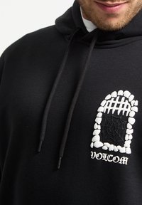 Hoodie noir avec un grand design graphique sur la poitrine, représentant une fenêtre encadrée de pierres avec une toile d'araignée, et le texte "VOLCOM".