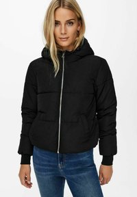 Doudoune noire avec capuche, fermeture éclair à l'avant, poignets élastiques et motif matelassé. Portée sur un jean bleu, avec une texture douce et lisse.