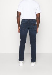 Jeans de denim azul oscuro con un ajuste Slim, con bolsillos traseros y una etiqueta de la marca. Llevados con una camiseta blanca y zapatillas blancas.
