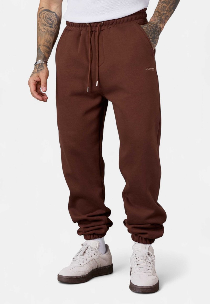 Braune Sweatpants mit elastischem Bund, Kordelzug, elastischen Bündchen und einer Seitentasche; kombiniert mit weißen Sneakers mit grauen Sohlen.