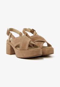 Suede beige block-heeled sandals featuring crisscross straps, an ankle buckle, and stitching detail. Textured surface with a soft finish.