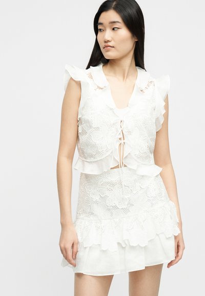 Γυναίκα που φοράει λευκό δαντελένιο αμάνικο τοπ με κορδόνια στο μπροστινό μέρος και ταυτόχρονη μίνι φούστα με βολάν, κοιτάζει πλάγια σε απλό φόντο.