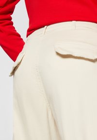 Beige, high-waisted trousers with a tailored fit, featuring two flap pockets on the back and a smooth texture. Smooth cotton material.