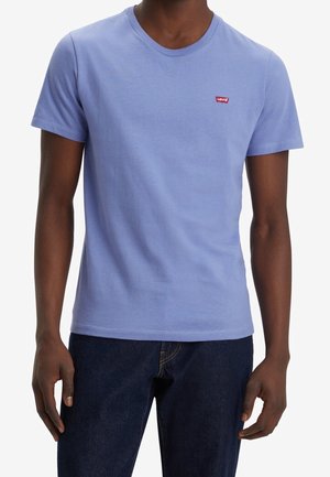 Mężczyzna w jasnoniebieskiej koszulce Levi’s i ciemnoniebieskich dżinsach na białym tle, ręce swobodnie opadające wzdłuż ciała.