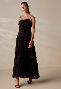 Juodas maxi sukneles su gėlių aplikacijomis, prigludusia liemene ir plonais petnešomis, turinčios erdvų sijoną ir sluoksniuotą tekstūrą. Atviros pirštinės kulniukai.