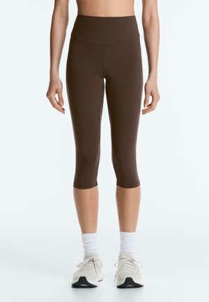 Nő, sötétbarna, magas derekú, rövid leggingsben, fehér zokniban és bézs futócipőben áll egy egyszínű világos háttér előtt.