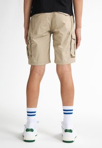 Shorts cargo beige con due tasche laterali, una vestibilità rilassata e un orlo poco sopra il ginocchio, abbinati a calze bianche con strisce blu e scarpe da ginnastica.