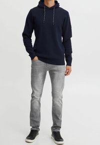 Homme portant un hoodie bleu marine, un jean gris clair et des baskets noires, se tenant contre un fond blanc uni.