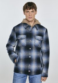 Giacca camicia a scacchi blu e nera con cappuccio tan, due tasche frontali, bottoni a scatto e una texture morbida.